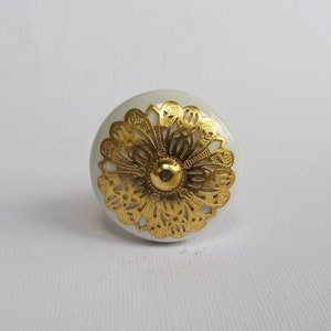 May include: A white ceramic cabinet knob with a gold filigree design. The knob has a rounded shape with a central gold accent. The gold filigree is in a floral pattern. This decorative hardware is suitable for furniture.