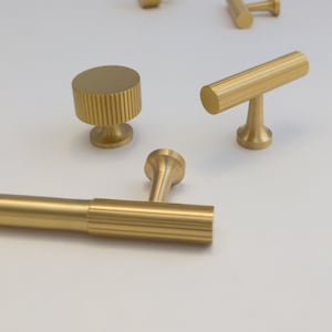 May include: A set of five gold-toned cabinet knobs and pulls. The set includes two round knobs, two rectangular pulls, and one T-shaped pull. All of the hardware features a ridged design.