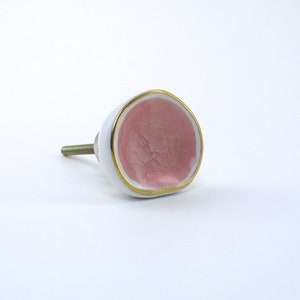 May include: A white ceramic cabinet knob with a pink interior and gold trim. The knob has a rounded shape and a metal screw for installation. This decorative hardware adds a touch of elegance to any furniture piece.