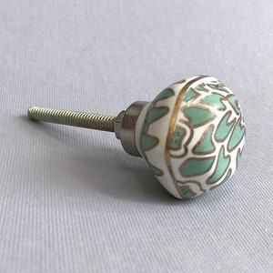 May include: A white ceramic cabinet knob with a green and gold floral design. The knob has a silver screw.