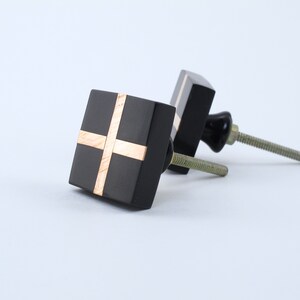 May include: Two black square cabinet knobs with gold trim. The knobs have a cross design and are attached to screws.