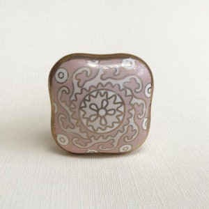 May include: A square cabinet knob with a pink background and a gold and white floral design. The knob has a gold-colored metal edge. This decorative hardware is suitable for drawers and cabinets.