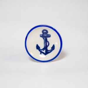 May include: A round, white ceramic knob with a blue anchor design in the center. The knob has a blue border and is set against a white background. Nautical-themed hardware.