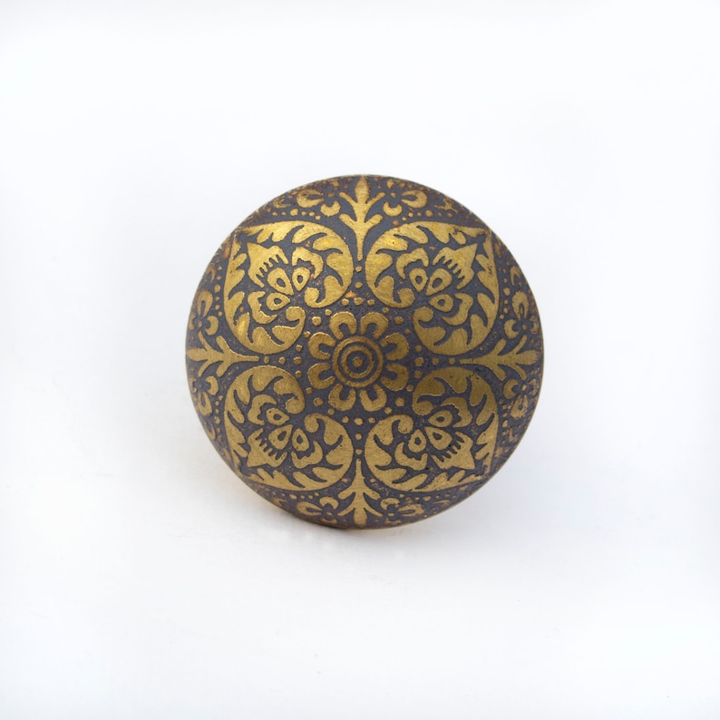 May include: A round, decorative knob with a dark gray base and intricate gold floral patterns. The design features a central flower surrounded by symmetrical leaf and petal motifs, creating a vintage aesthetic. The knob is likely made of metal.