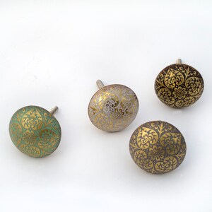 May include: Four decorative cabinet knobs with ornate gold patterns on a variety of colored backgrounds, including teal, gold, and gray. Each knob has a silver screw for attachment. The knobs are on a white background.
