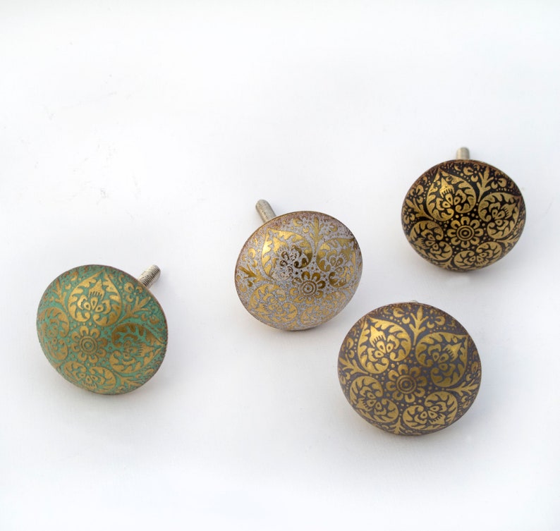 May include: Four decorative cabinet knobs with ornate gold patterns on a variety of colored backgrounds, including teal, gold, and gray. Each knob has a silver screw for attachment. The knobs are on a white background.