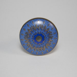 May include: A round cabinet knob with a blue and gold floral design. The knob has a brass-colored rim and a central blue circle. The design features gold-colored, stylized floral patterns on a blue background.