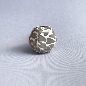 May include: A small, white ceramic vase with a unique, rounded shape. The vase is decorated with a gray floral pattern and gold accents. The vase is a decorative item.