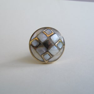 May include: A round, white, and gold decorative knob with a geometric pattern. The knob is made of mother of pearl and has a gold trim.