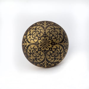 May include: A round, decorative object with a dark brown base and an intricate gold floral pattern. The design features a central flower surrounded by symmetrical, stylized floral motifs. The object is set against a white background.