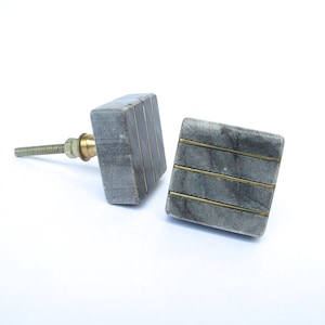 May include: Two square cabinet knobs. The knobs are gray with gold-colored metal accents. One knob has a threaded screw and brass hardware. The other knob is positioned next to it. The knobs are made of a stone material.