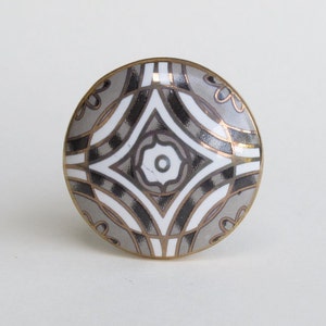 May include: A round ceramic knob with a geometric pattern in shades of gray, black, and gold. The knob has a white center with a smaller circle in the middle.
