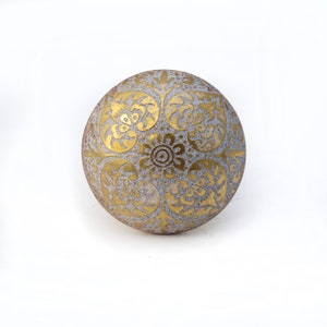 May include: A round, gray decorative knob with a gold floral pattern. The knob features a central flower design surrounded by ornate, swirling leaf and petal motifs. The knob is likely made of ceramic or stone.