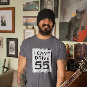May include: A man wearing a grey t-shirt with a white graphic of a road sign that says "I can't drive 55". He is holding a brown and black electric guitar.
