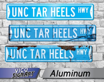 May include: Three blue highway signs with white text that reads "UNC TAR HEELS HWY". The signs are made of aluminum and are mounted on a brick wall. The bottom sign has rust.
