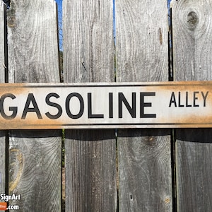 May include: A rectangular wooden sign with the words "GASOLINE ALLEY" in black letters. The sign has a white background with a distressed, aged appearance and is mounted on a weathered wooden fence.