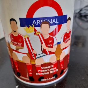 May include: A white ceramic mug featuring the Arsenal F.C. Premier League Champions 2025/26. The mug has a red and white circular design with the word "ARSENAL" in blue. Illustrations of players in red and white uniforms are also featured.