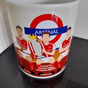 May include: White ceramic mug featuring the Arsenal logo and illustrations of players holding a trophy. The mug includes the text "Arsenal F.C. Premier League Champions 2020/26".