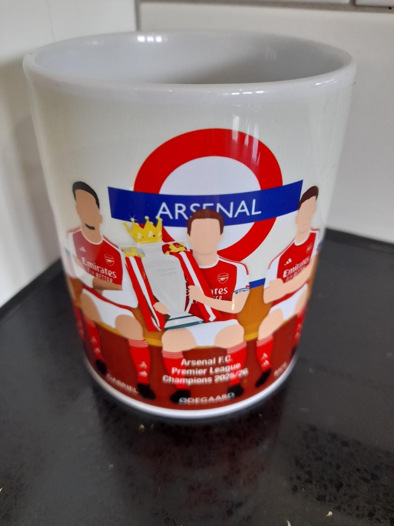 May include: White ceramic mug featuring the Arsenal logo and illustrations of players holding a trophy. The mug includes the text "Arsenal F.C. Premier League Champions 2020/26".