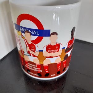 May include: White ceramic mug featuring an Arsenal design. The mug has a red and blue circular logo with the word "ARSENAL". It also displays illustrations of Arsenal players in their red and white uniforms. Text on the mug reads "Arsenal F.C. Premier League Champions 2025/26".
