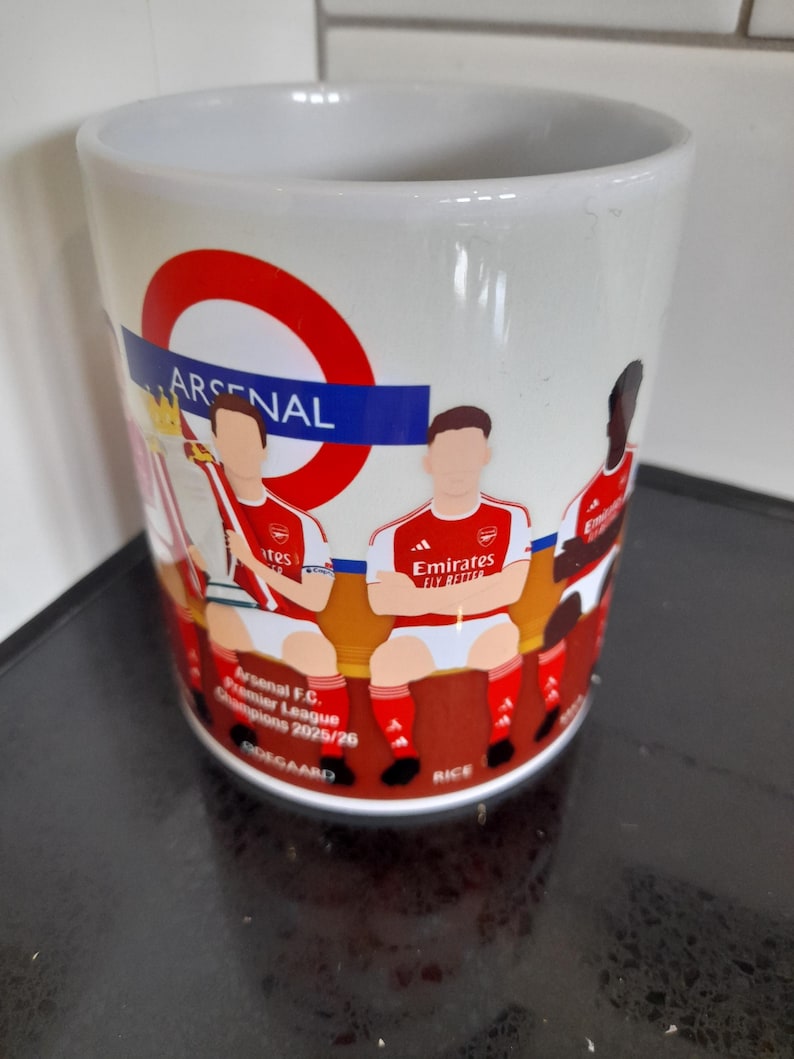 May include: White ceramic mug featuring an Arsenal design. The mug has a red and blue circular logo with the word "ARSENAL". It also displays illustrations of Arsenal players in their red and white uniforms. Text on the mug reads "Arsenal F.C. Premier League Champions 2025/26".