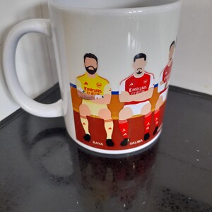 May include: White ceramic mug featuring a colorful illustration of soccer players in team uniforms. The mug has a curved handle and the names "Raya" and "Saliba" are visible. The mug is likely a fan item for a soccer team.