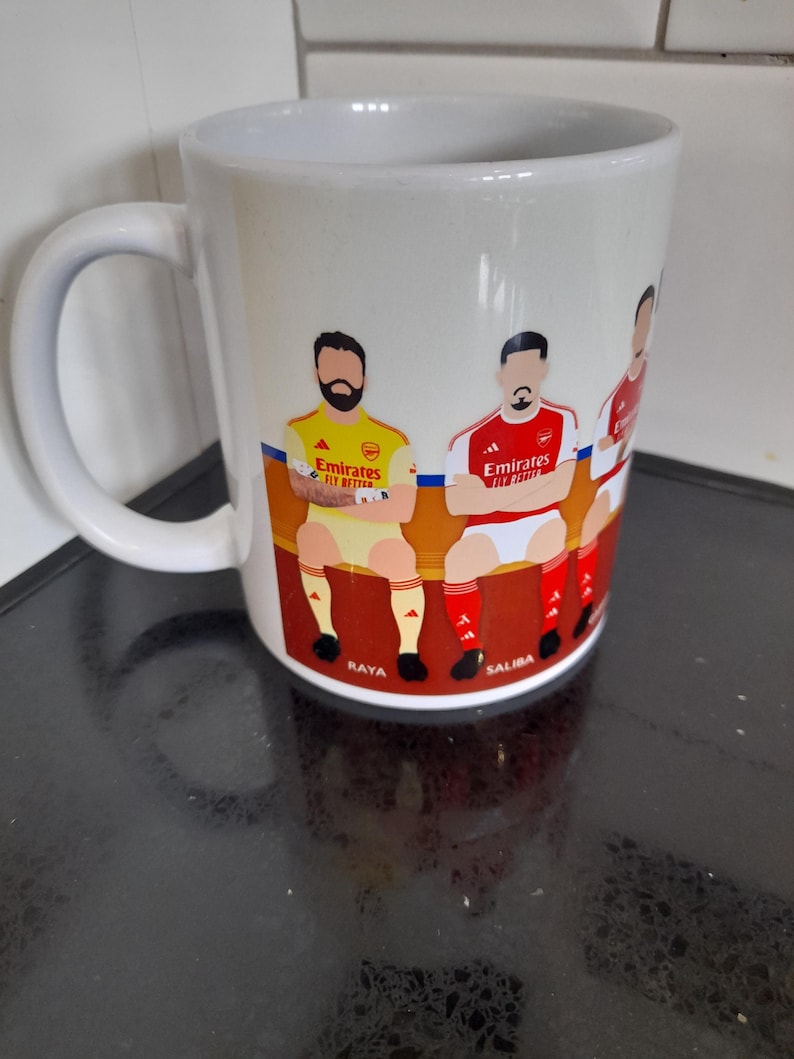 May include: White ceramic mug featuring a colorful illustration of soccer players in team uniforms. The mug has a curved handle and the names "Raya" and "Saliba" are visible. The mug is likely a fan item for a soccer team.
