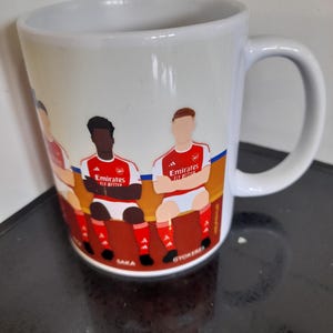 May include: White ceramic mug featuring a colorful illustration of soccer players in red and white uniforms. The mug has a curved handle and the text "Emirates Fly Better" is visible on the design. The names "Saka" and "Gyokeres" are also visible.