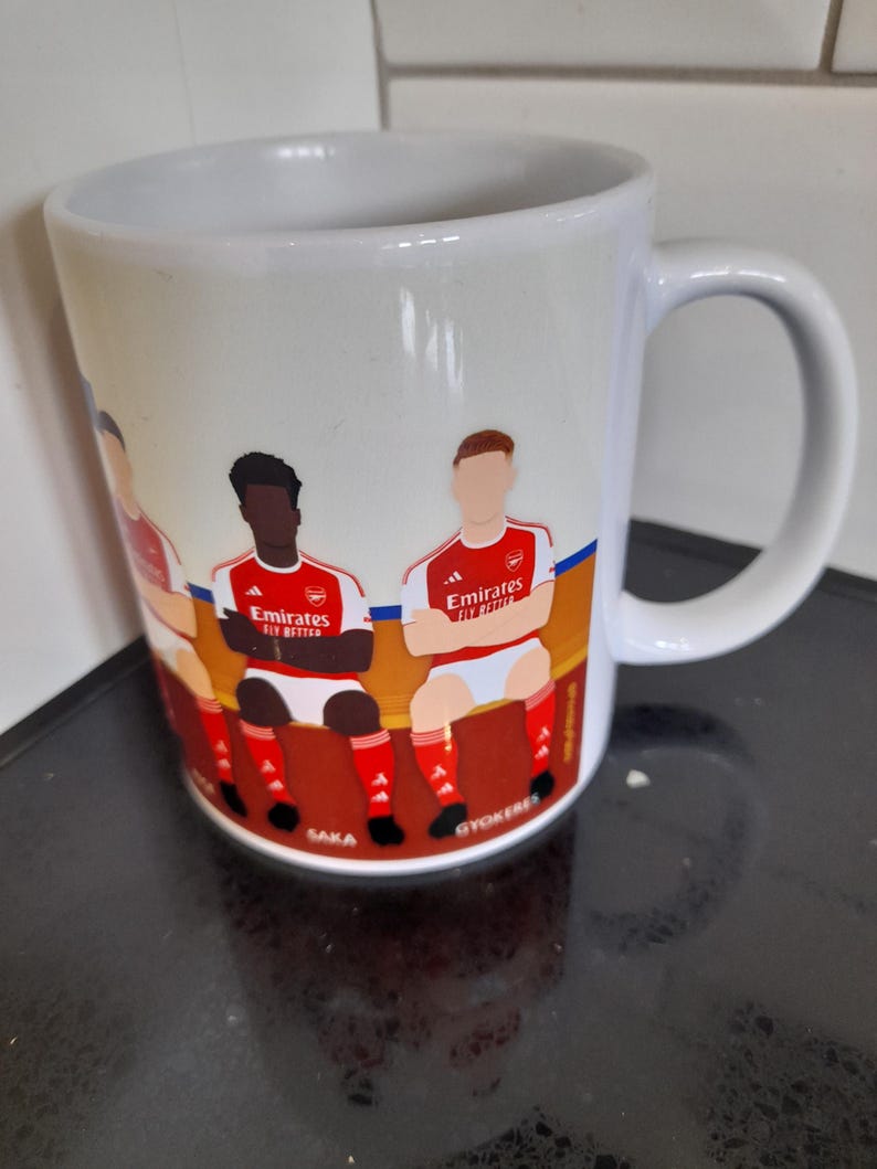May include: White ceramic mug featuring a colorful illustration of soccer players in red and white uniforms. The mug has a curved handle and the text "Emirates Fly Better" is visible on the design. The names "Saka" and "Gyokeres" are also visible.