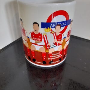May include: White ceramic mug featuring a design of the Arsenal football team. The mug has a red and white color scheme with the Arsenal logo and text. The design includes illustrations of players and the Premier League trophy.