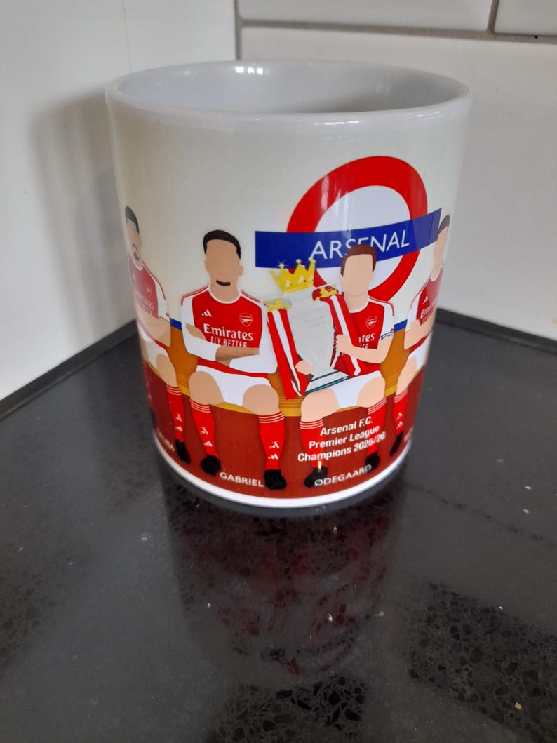 May include: White ceramic mug featuring a design of the Arsenal football team. The mug has a red and white color scheme with the Arsenal logo and text. The design includes illustrations of players and the Premier League trophy.