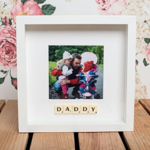 May include: White picture frame with a photo of a father and two young children. The photo is surrounded by a white mount and the word "DADDY" is spelled out in Scrabble tiles below the photo.