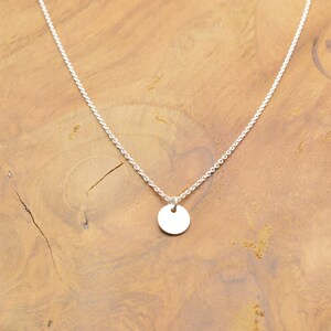 Könnte beinhalten: Eine silberne Kettenkette mit einem kleinen, runden silbernen Anhänger. Die Kette liegt auf einer Holzoberfläche mit den Worten "Hand Made", die in das Holz eingeprägt sind.