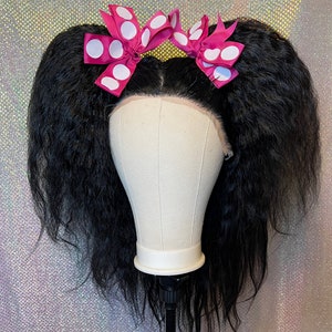 Może przedstawiać: Czarna peruka z dwoma różowymi kokardkami w białe kropki na białej głowie manekina.