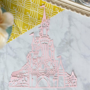May include: A pink paper cutout of a fairytale castle with intricate details. The castle has multiple towers, windows, and a large arched entrance.