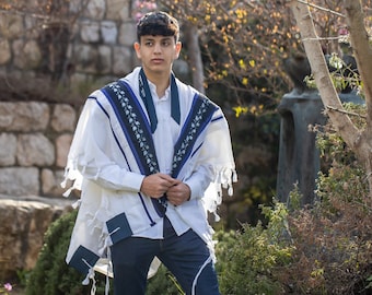 Puede incluir: Un chal de oración tallit blanco con bandas decorativas azul oscuro y negras con un patrón floral, usado sobre una camisa blanca de botones y jeans azul oscuro. El chal tiene flecos anudados en los bordes y las esquinas.