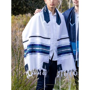 May include: White prayer shawl with blue and silver stripes and tassels. The shawl has a dark blue collar and corner accents. The prayer shawl is worn over a white shirt and dark pants. The image is taken outdoors.