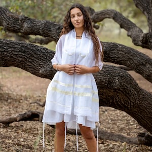 May include: A white sleeveless dress paired with a matching white tallit shawl featuring gold-toned horizontal stripes and white tzitzit fringes at the corners. The ensemble is completed with white flat shoes, creating a coordinated monochromatic look.