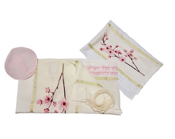 Puede incluir: Un conjunto de artículos de oración color crema, que incluye una kipá, un talit y una bolsa a juego. El talit y la bolsa presentan un borde dorado y un delicado diseño de flor de cerezo rosa. Se ve texto hebreo en la bolsa.