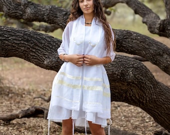 May include: A white sleeveless dress paired with a matching white sheer shawl featuring gold horizontal stripes. The shawl has decorative embroidery along the neckline and long white fringe tassels hanging from the bottom corners of the garment.