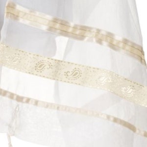 May include: A white sheer fabric prayer shawl featuring horizontal bands of cream-coloured ribbon and a central decorative trim adorned with a repeating paisley pattern. The lightweight material has a translucent quality with visible woven textures.