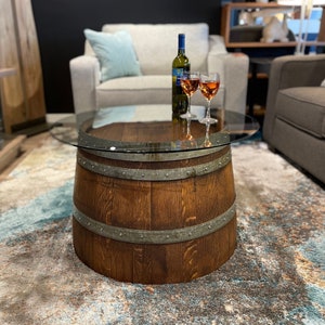 May include: A wooden barrel coffee table with a glass top. The table is in a living room setting with a couch and a chair. There are two glasses of wine and a bottle of wine on the table.