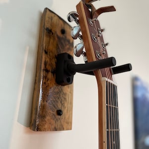 May include: A wooden wall mount guitar hanger with a black metal hook holding an acoustic guitar with a natural wood finish.