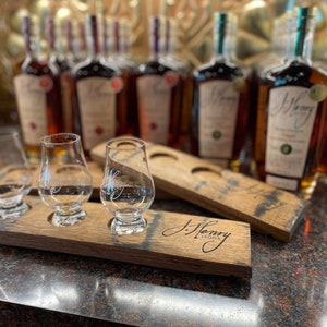Może przedstawiać: Trzy przezroczyste szklane kieliszki do degustacji znajdują się na drewnianej tacy do degustacji z wygrawerowanym napisem "J. Henry & Sons" z przodu. Taca znajduje się przed rzędem bursztynowych butelek z zielonymi korkami.