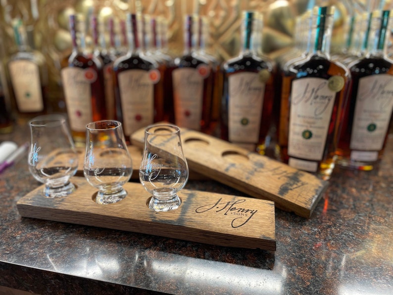Może przedstawiać: Trzy przezroczyste szklane kieliszki do degustacji znajdują się na drewnianej tacy do degustacji z wygrawerowanym napisem "J. Henry & Sons" z przodu. Taca znajduje się przed rzędem bursztynowych butelek z zielonymi korkami.