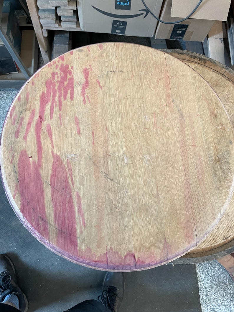 May include: A close-up of a round wooden barrel top with a reddish-brown stain. The wood is light brown and has a natural grain pattern.