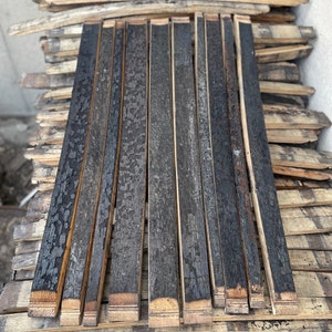 May include: A stack of charred oak barrel staves, with a dark, textured surface. The staves are arranged in a neat pile, with some showing the natural wood grain. The ends of the staves have a lighter, unfinished appearance. The image suggests a rustic, reclaimed wood theme.