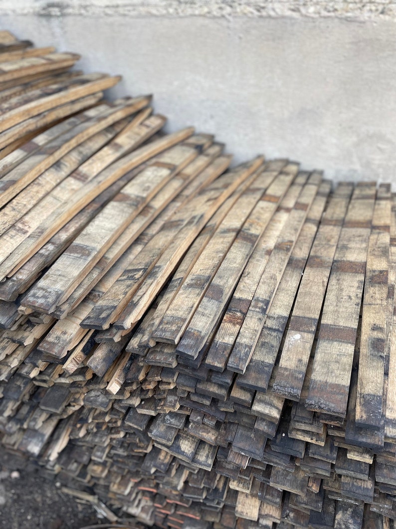 May include: A stack of weathered, curved wooden barrel staves. The staves are various shades of brown and gray, with dark charring on some edges. The wood grain is visible, suggesting a rustic or reclaimed aesthetic.