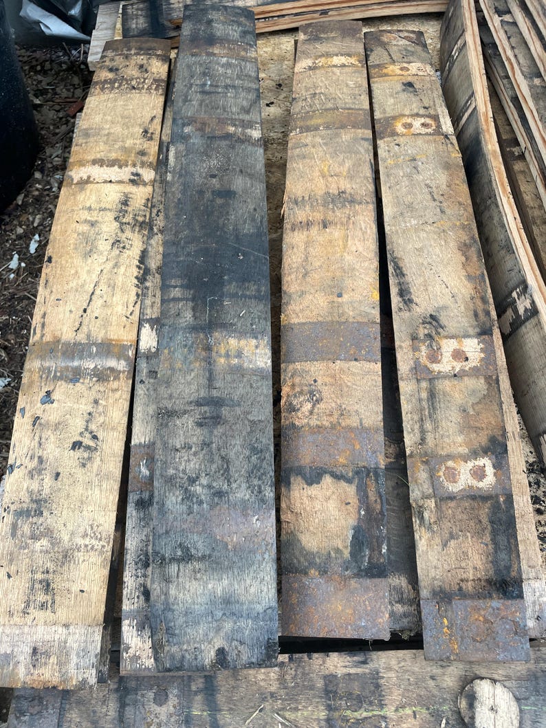May include: Four reclaimed wooden barrel staves, showcasing a rustic aesthetic. The staves vary in color, from light tan to dark charcoal, with visible grain and metal bands. These pieces are ideal for woodworking projects or adding a vintage touch to decor.
