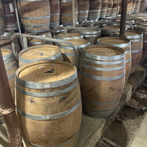 May include: A row of wooden barrels stacked in a winery. The barrels are made of oak and have metal bands around them. The barrels are labelled with the name "Justin Vineyards & Winery".